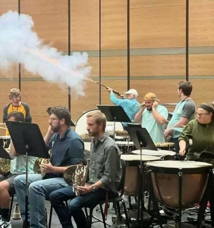 Orchestra practice scene with musicians unfazed by a man creating smoke with a long stick, capturing a 'hmmm' moment.