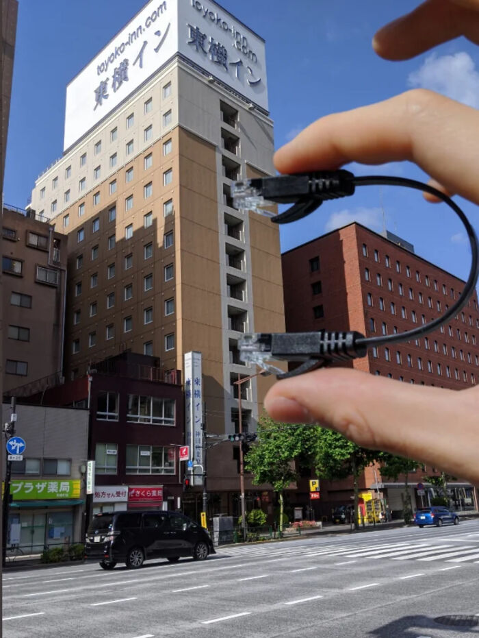 Fingers holding cable above a city street, resembling a giant plug for the building, creating a quirky optical illusion.