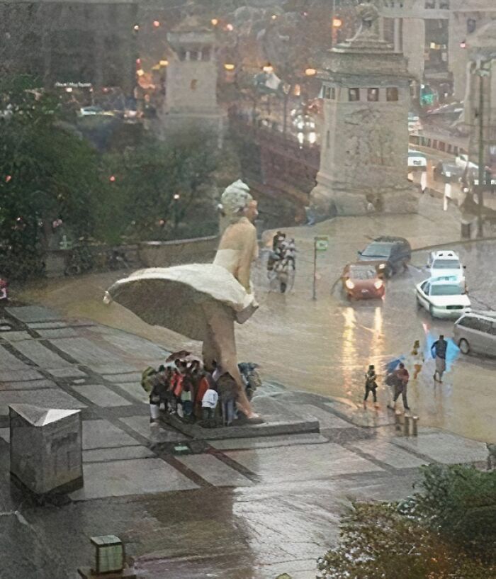 Group shelters under large statue's dress during rain, a quirky sight making them go 'hmmm'.